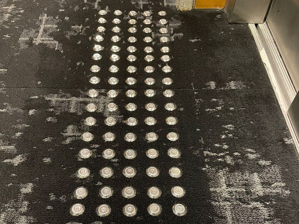 Piso tátil de inox em formato de alerta instalado sobre carpete, utilizado para orientação de pessoas com deficiência visual próximo ao elevador.