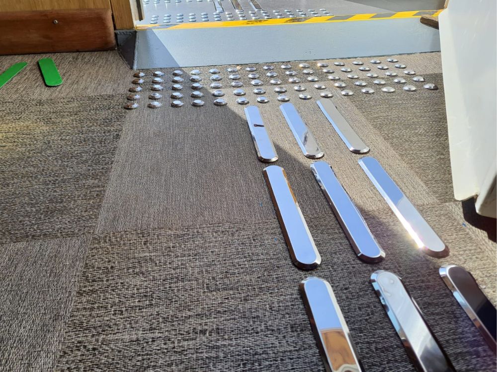 Detalhe da instalação de piso tátil inox em carpete, com elementos de alerta e direcionais fixados com precisão em entrada acessível.