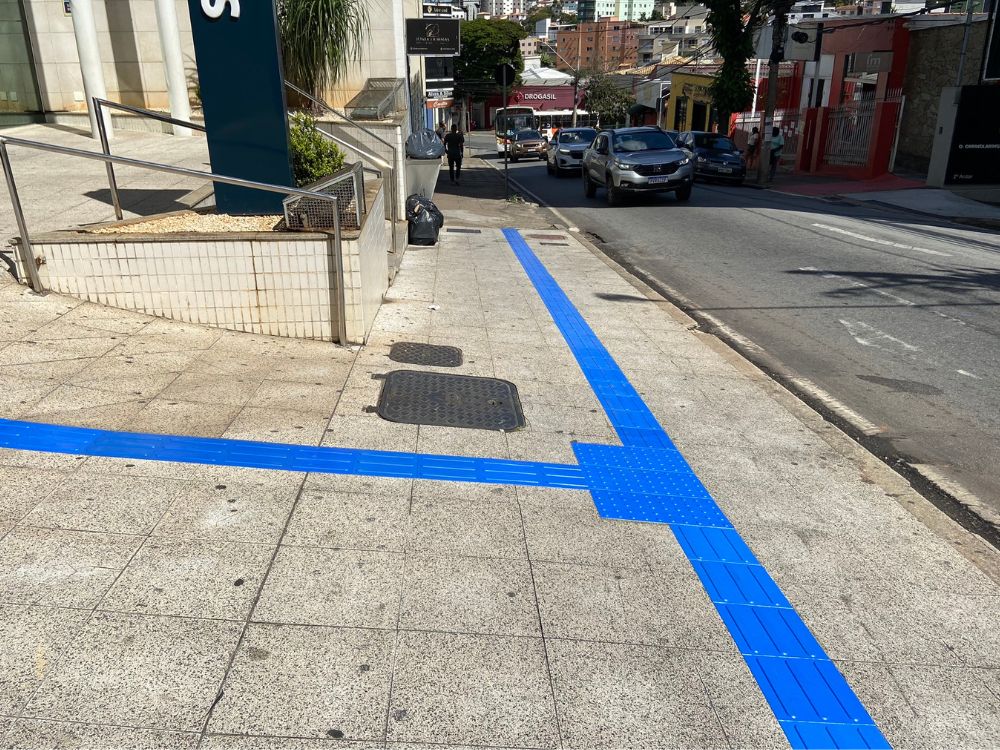 Piso tátil azul instalado em calçada urbana, com alerta posicionado a 50 cm do meio fio, conforme norma técnica de acessibilidade para travessia segura.