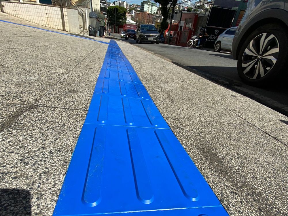 Vista em perspectiva do piso tátil direcional azul conduzindo até a região próxima ao meio fio, respeitando a distância adequada de segurança.
