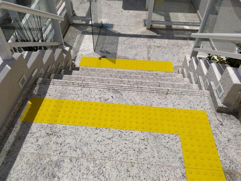 Escada com piso tátil amarelo em PVC aplicado no topo e base, seguindo a NBR para garantir acessibilidade e segurança a pessoas com deficiência visual.