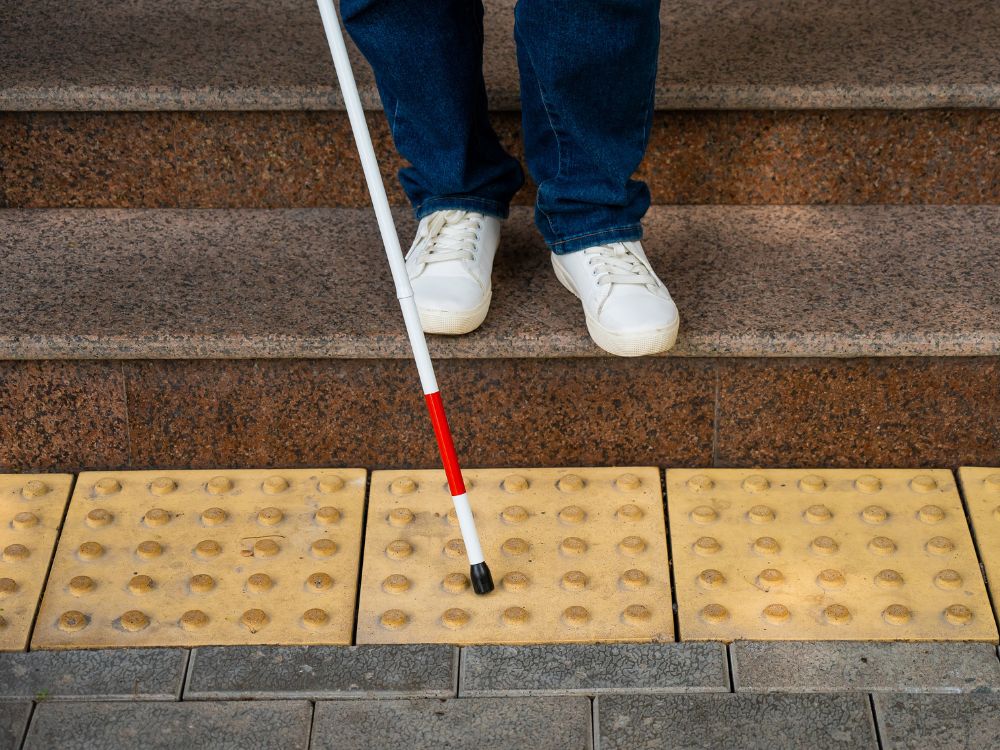 Os pés e a bengala de uma pessoa com deficiência visual sobre um piso tátil amarelo, na base de uma escada. A bengala e o piso tátil são ferramentas essenciais para a acessibilidade para cegos.