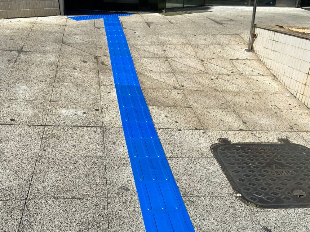 Uma passarela com piso de concreto e piso tátil direcional azul, que possui relevos longitudinais para guiar a pessoa, ilustrando um exemplo de como colocar piso tátil na calçada.