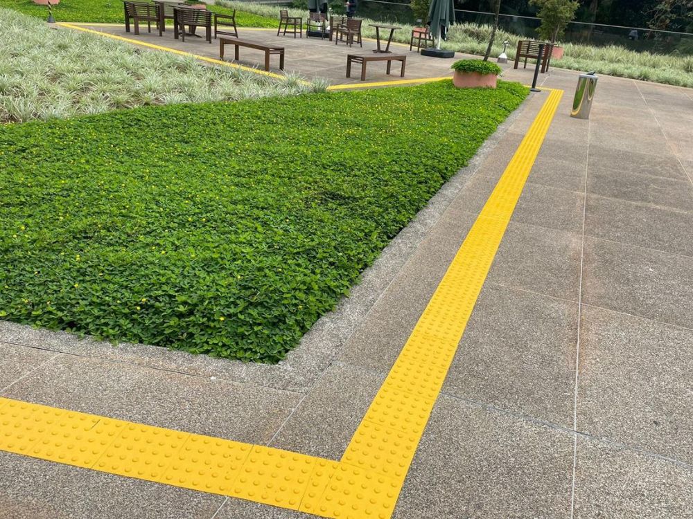 Uma calçada pública com um piso tátil de alerta amarelo, com relevos circulares. A instalação do piso tátil demonstra a necessidade de saber como colocar piso tátil na calçada para garantir a segurança dos pedestres.