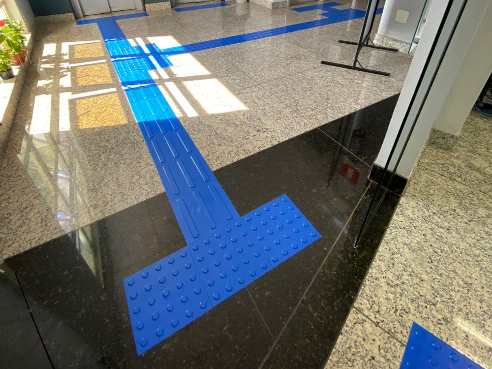 Piso tátil azul com faixas direcionais e de alerta aplicado sobre granito, exemplificando sinalização para deficientes visuais em corredores internos.
