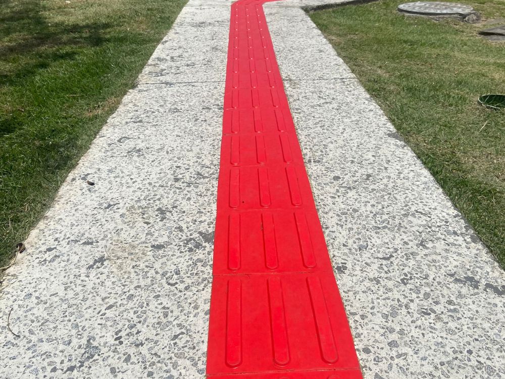 Piso tátil em rampa com faixa direcional vermelha aplicada em calçada, facilitando o deslocamento e a identificação de percurso acessível
