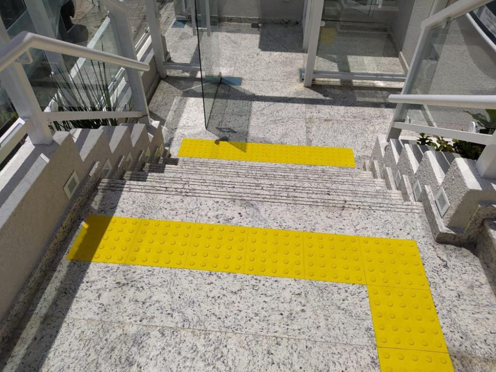 Piso tátil amarelo em escada com sinalização de alerta, evidenciando o tamanho do piso tatil adequado para segurança de pessoas com deficiência visual.