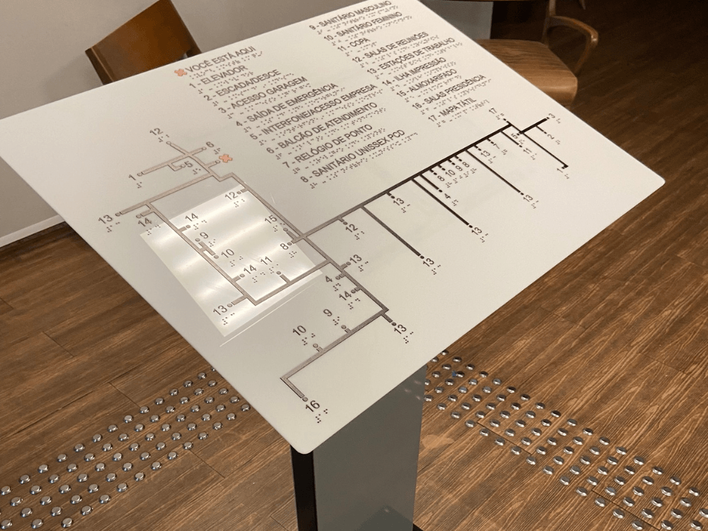Mapa tátil instalado em um suporte apresenta a planta de um edifício com inscrições em relevo e piso tátil ao redor.
